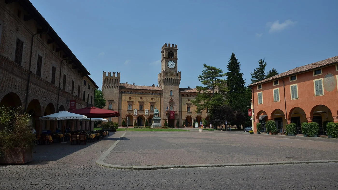 Ein weiter gepflasterter Platz in Busseto, gesäumt von Arkaden und Café-Sonnenschirmen, mit einem Uhrturm am anderen Ende unter blauem Himmel