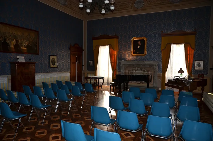 A music salon with blue damask walls, a grand piano, a portrait in a gilt frame and rows of blue chairs on a patterned wooden floor