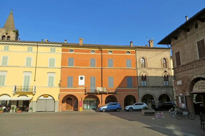 Case gialle e arancioni color pastello con portici su una piazza di Busseto, con auto parcheggiate e biciclette sotto un cielo azzurro