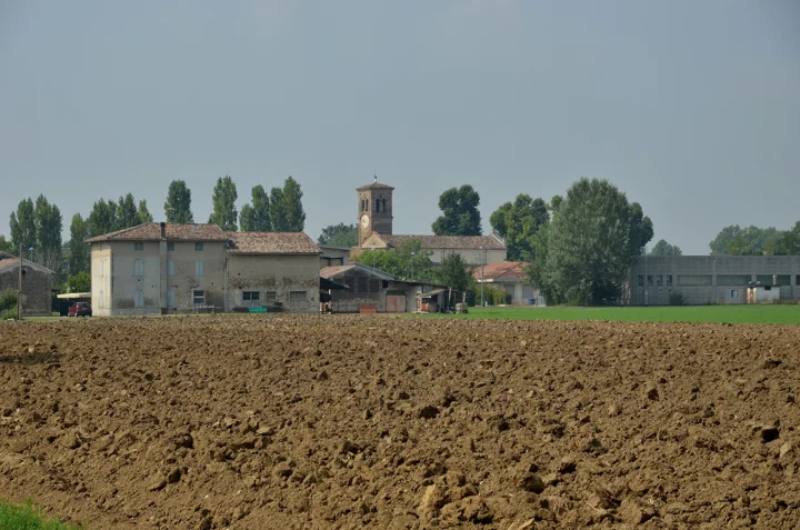 Campi arati pianeggianti con bassi edifici rurali, pioppi e un campanile lontano sotto un cielo velato