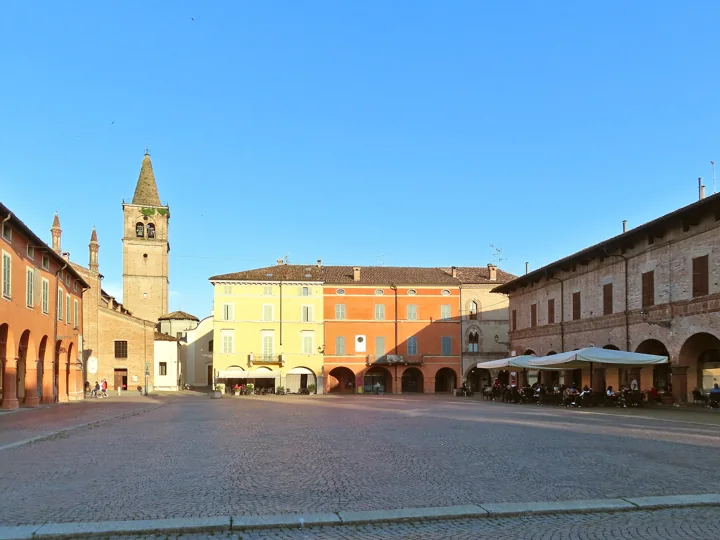 Ein weiter gepflasterter Platz in Busseto mit pastellgelben und orangefarbenen Palazzi, einem Glockenturm aus Backstein und Café-Sonnenschirmen unter Arkaden