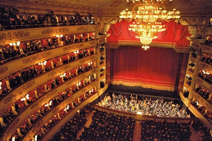 The red and gold auditorium of La Scala in Milan seen from a high box, with tiers of boxes, a great chandelier and an orchestra on stage
