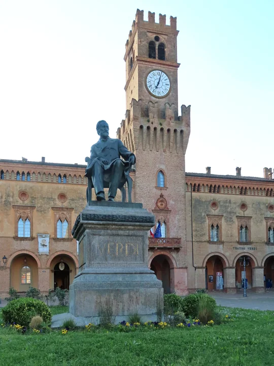 A bronze statue of Verdi seated on a tall stone pedestal in front of a brick castle with a clock tower
