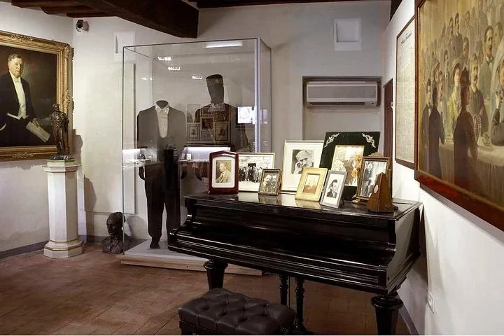 A museum room with a black piano, framed photographs, a portrait and display cases of clothes and mementos