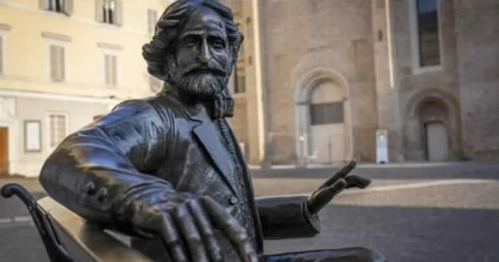 A bronze figure of Verdi sitting on a bench with one hand raised, in front of pale town buildings