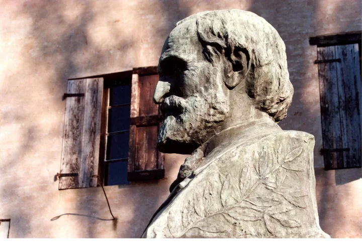A pale weathered bust of the old Verdi with a full beard in front of the birthplace's wooden shutters