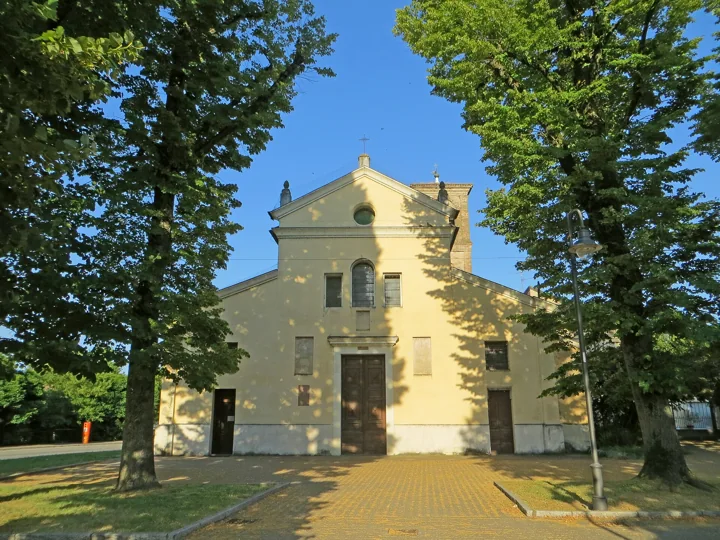 The pale facade of the church of San Michele Arcangelo at Roncole between two tall trees, under a blue sky