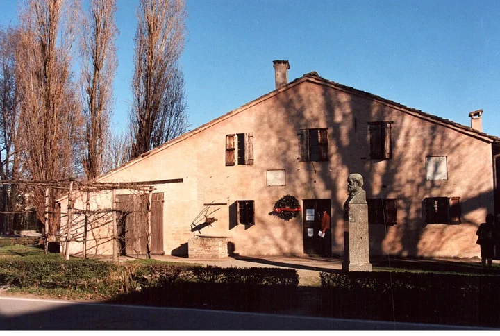 La casa natale di Verdi a Roncole nel sole basso, con pioppi spogli dietro, un pergolato di vite e un busto accanto alla porta