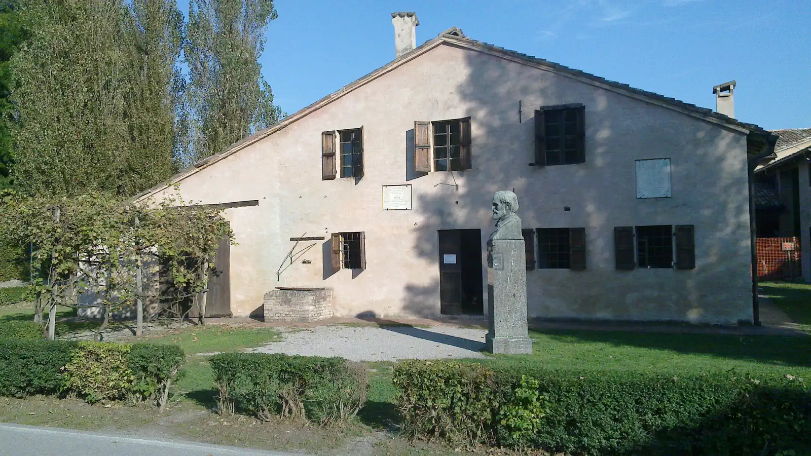 The pale plastered birthplace of Verdi at Roncole with shuttered windows, a pergola and a bust on a pillar in the garden, under a blue sky